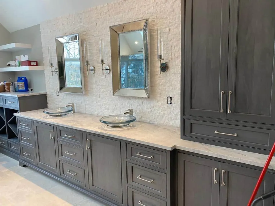 Long gray vanity with a white veined stone top and two glass vessel sinks in front of a white stacked-stone wall