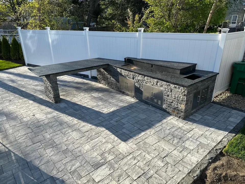 L-shaped outdoor kitchen with a dark stone top and a raised bar on a stacked-stone base, on a paver patio